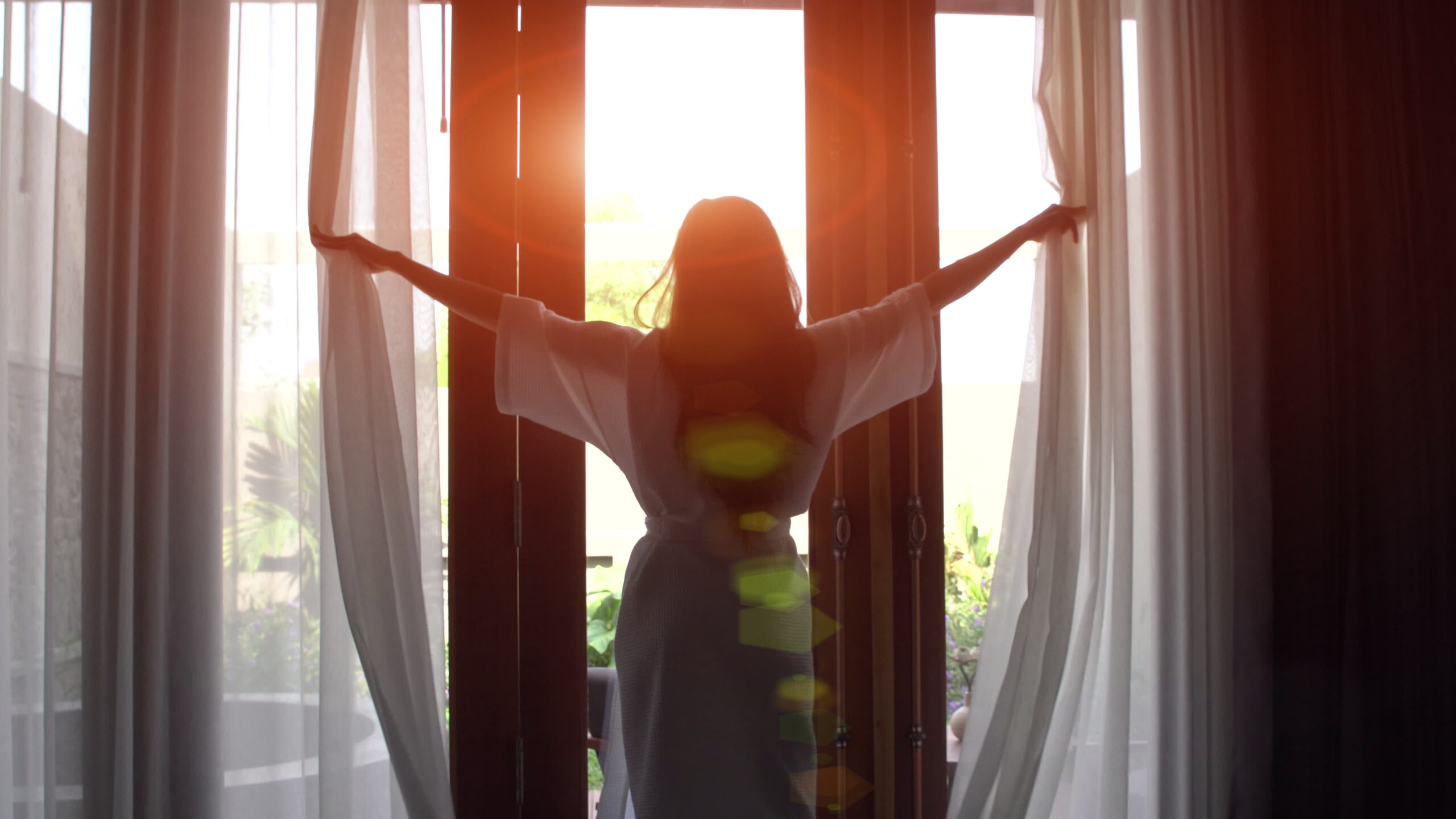 Young woman in bathrobe open curtains and stretch standing near the window at home.